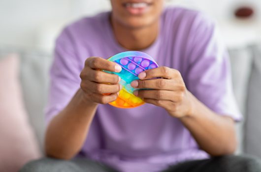 Closeup view of teenager playing with antistress POP IT toy at home. Unrecognizable black youth using occupational therapy toys to relieve stress or relax indoors