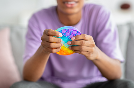 Closeup view of teenager playing with antistress POP IT toy at home. Unrecognizable black youth using sensory plaything to relieve stress or relax indoors