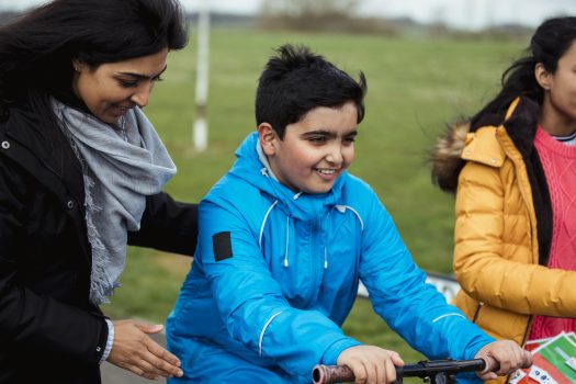 A mother holding her son, supporting him on his scooter, with his sister walking by his side.