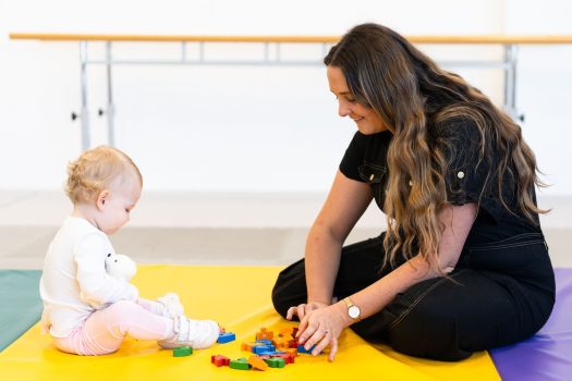 Woman sitting crosslegged on the ground, playing with a child on a playmat in a therapy room